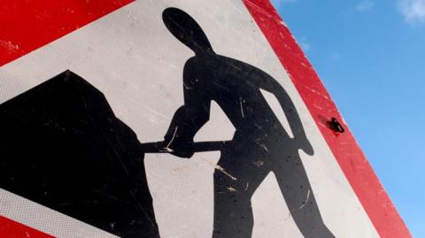 Close-up of a UK road work sign. It is triangular in shape with a red outer rim while the detail of the sign shows a lone workman digging a shovel. The sign is set against a blue sky.