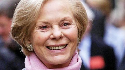 An older woman with a tidy blond bob, silver earrings and pink scarf. She is looking just off-camera and smiling.