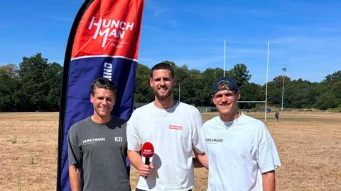 A group of three brothers stood next to each other. One, stood in the middle, is holding a microphone.