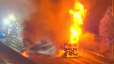 A large fire on a road, showing a lorry in flames and firefighters working to put it out. Fire engines are on the road and you can only just see their lights.