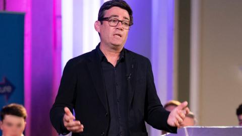 Andy Burnham speaking at an event wearing a black shirt and jacket