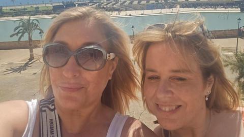 Two women - one wearing sunglasses, the other with sunglasses on her head - smiling on a sunny day. A palm tree stands in the background.