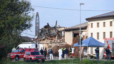 A building reduced to rubble with emergency services cars parked in front of it