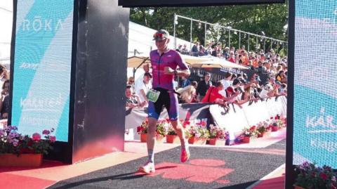 Braden Morris passing over the finish line. He is wearing blue and pink running gear and a cap with sunglasses.