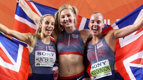 (L-R) Georgia Hunter Bell, Molly Caudery and Keely Hodgkinson pose for a photo at the World Athletics Indoor Championships. They're holding union jack flags behind them and wearing Team GB athletic wear