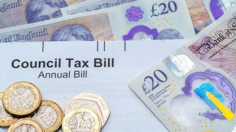 A number of £20 notes are placed on a flat surface, with a council tax bill showing in the middle. One pound and 50p coins are also overlayed.