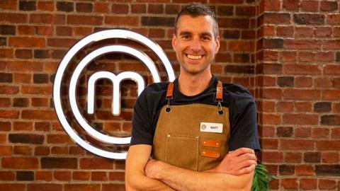 Matt, who has dark hair and is wearing a brown apron, smiles for a photo in the MasterChef studio.