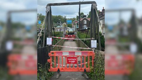 A suspension bridge with a 'footpath closed' sign and barrier in front of it. The bridge has red and white tape on it as well as some white signs. On the opposite side of the bridge is some scaffolding.