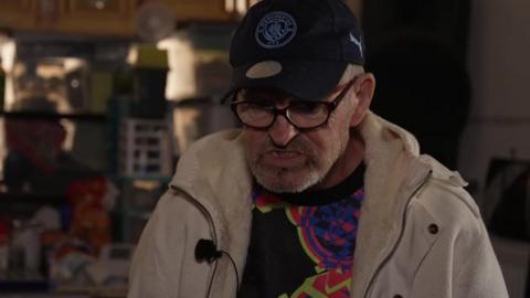 Lawrence Cook, wearing a black cap, beige coat and black-rimmed glasses, looks at the floor in his darkened flat