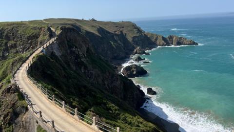 A coastal cliffside footpath curves along rugged headlands above turquoise water, with waves breaking against rocky outcrops below under a clear blue sky.