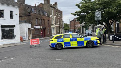 A police car is parked across a junction which has been cordoned off using white and blue police tape. Two police officers and a man are standing on the right talking. In the middle of the road there is a road closed sign.