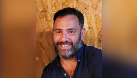James O'Connor is looking into the camera and smiling. He's wearing a navy shirt and he has brown hair. There's a bare wooden wall behind him.