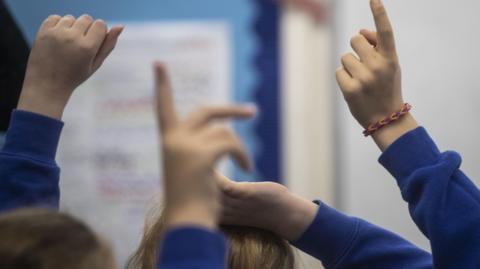 A primary school classroom with pupils putting up their hands. They have bright blue jumpers on.