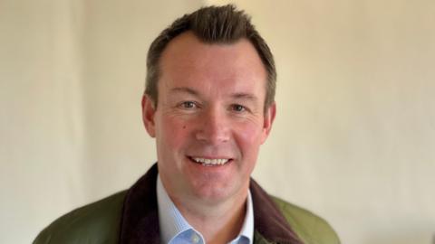 A man with brown hair is smiling at the camera. He is wearing a white and blue checked shirt with a green jacket that has a brown collar