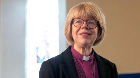 The Most Reverend Dame Sarah Mullally stands in a room looking away from the camera. She has bobbed blonde hair and is wearing a pink shirt with a clerical collar, black blazer and glasses. 