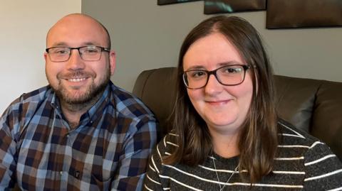 A balding man with a beard and glasses sitting on a sofa at home next to a woman with long brown hair and glasses 