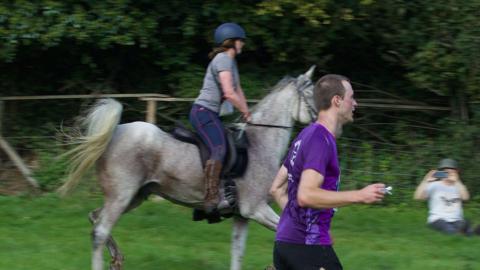 A man in a purple T-shirt runs through a field. Behind him, a woman rides a horse which also appears to be galloping. A spectator holding a phone up to take a picture can be seen in the background. 
