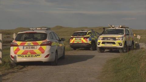 Three Irish police cars are parked at the entrance to a beach. Two face away from the camera and towards the sea while a third, on the right of the other two, faces forwards. They are parked on a gravel path that splits sand dunes