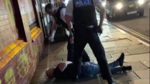 Two police officers in uniform stands over Owen Andrews, wearing a black t-shirt and jeans, who is lying on a pavement on his back. One officer leans forward with his arm out-stretched and a jet of white spray stretches from a canister in his hand to Mr Andrews' face.