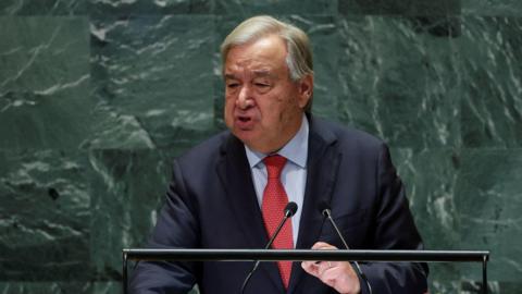 UN Secretary General Antonio Guterres giving a speech at the United Nations in New York. He is standing against the distinctive green marbled backdrop at a lectern, wearing a red tie, dark suit jacket and blue shirt