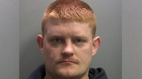Mugshot of Brian Light. He has short bright ginger hair and has a small ginger moustache. He has blue eyes and is wearing a navy hoodie. 