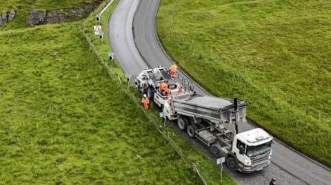 A lorry lowers chippings on to a road while workman sit on the back of the vehicle. The scene - a rural road with sweeping bend and green fields on either side - is pictured from above. The lorry has a white cab, in the foreground, and the trailer behind is hydraulic and tilted to about 25 degrees to allow the grey coloured contents to pour into other heavy equipment behind.