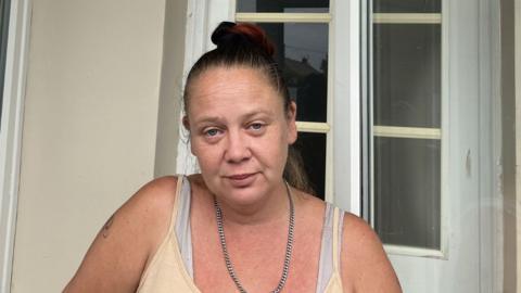 Head and shoulders image of Laura Smith who has dark hair tied in a bun outside a house in front of a white-framed glass door, wearing a light-coloured vest top and chain necklace. 