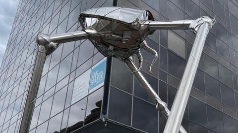 A shiny insect-like metal structure on three cylindrical legs stands in front of a tall glass-fronted building. There are two winding tentacles coming out the back of the statue.