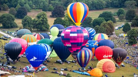 About two dozen hot air balloons take off from Ashton Court. Crowds can be seen standing around the balloons as some of them begin to leave the ground.
