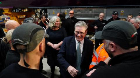 Sir Keir Starmer smiling as he meets workers at the Hitachi factory. He is wearing glasses, a navy suit, white shirt and a tie. Two workers in the foreground are facing away from the camera. They are wearing dark tops and green baseball caps.