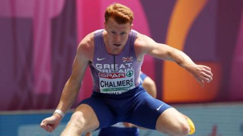 Alastair Chalmers competing in the hurdles. He is wearing a Great Britain vest and blue shorts.