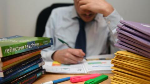 Stock image of a teacher marking at his desk. There are a pile of exercise books and reference books. The teacher's image is blurred