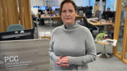 Alison Hernandez has brown hair pulled back from her face. She is wearing a grey jumper and standing near a sign which reads PCC Office of the Police and Crime Commissioner for Devon and Cornwall.