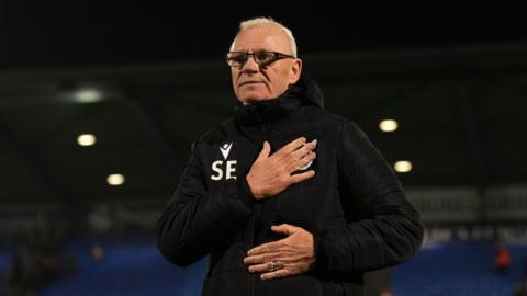 Steve Evans stands on the pitch with one hand across his heart and one on his stomach