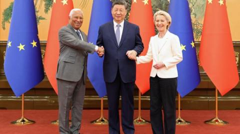 Chinese President Xi Jinping (centre) shakes hands with European Council President Antonio Costa and European Commission President Ursula von der Leyen at the Great Hall of the People in Beijing on 24 July, 2025. Behind them is a row of EU and China flags.