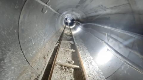 A view down an underground tunnel that is lit with bright white artificial light. There is a ladder track on the floor and various pipes and wires along the concrete walls.