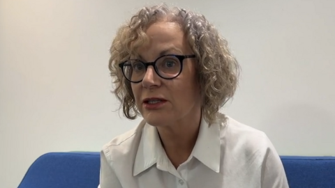 Jersey's children's commissioner Dr Carmel Corrigan. She has curly grey and blonde hair. She is wearing round black-rimmed glasses. She has a white blouse on and is sat on a blue sofa.