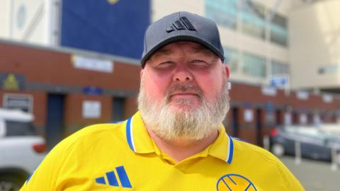 A man with a grey beard and a black cap wears a yellow Leeds United football club top. Behind him Elland Road stand is blurred.