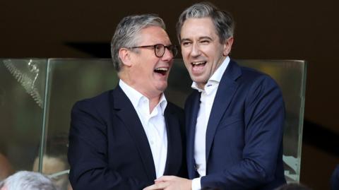 Sir Keir Starmer and Simon Harris. They are both smiling in celebration. Starmer has grey hair, wearing a dark blazer and white shirt. Harris has grey hair, wearing a navy blazer and white shirt.