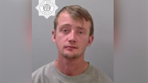 A man with short brown hair swept to one side. He has short facial hair and is wearing a grey sweatshirt.