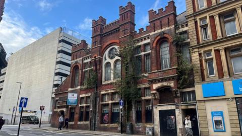 A red building with turret-like bricks at the top of the facade, alongside glass windows in the middle of the building.