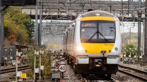 A yellow and silver train running on a railway track on a grey overcast autumnal day. Two members of staff sit in the front of the cabin, one is driving the train.