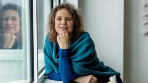 A woman posing in front of a window. She is leaning onto the window sill and resting her chin on knuckles. She has light brown, curly hair and is wearing a teal shawl over a long sleeve blue jumper. She is smiling into the camera.