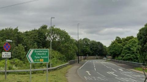 A green sign for the A19 on the A690 near Sunderland. Herrington Interchange is on the right. It is a section of a large, traffic light-controlled roundabout with trees in its centre.