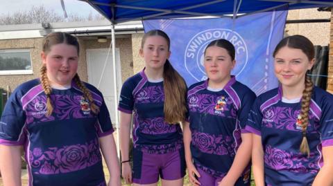 Four girls in their Swindon Roses purple rugby tops. They are smiling at the camera and standing in front of the a Swindon RFC banner. 