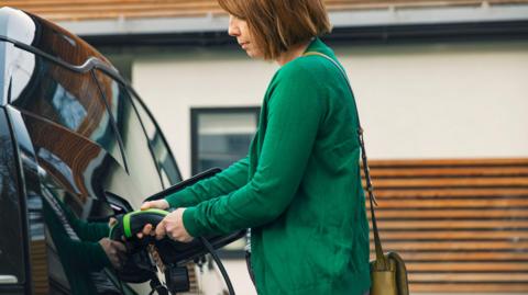 Woman wearing green cardigan charges black electric car outside of her home