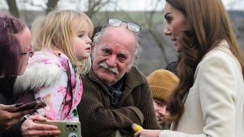 Catherine, Princess of Wales, with toy banana in hand, and watched by onlookers, chats to Lily-Rose after the unscheduled stop outside the Corgi factory in Ammanford