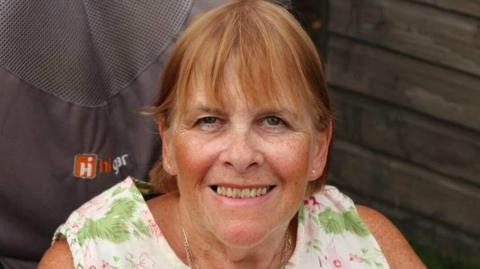 Linda Ashley-Davidson has a fringe and short hair. She is wearing a floral short sleeved dress and is sitting on a chair in front of a wooden wall.