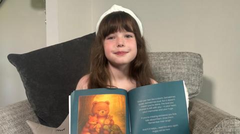 Daisy Blu Mathison holding her book open. She is sitting on a sofa and has long brown hair and is wearing a white headband. The book is open on a page displaying a cartoon bear, fox, tortoise and two rabbits all hugging.