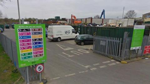 The entrance to Dorchester tip. The site is surrounded by steel fencing. There is a 5-miles-per-hour sign at the entrance. Inside the gates, several vehicles are lined up in front of a row of large dark green open-top shipping containers containing different types of rubbish and recycling.